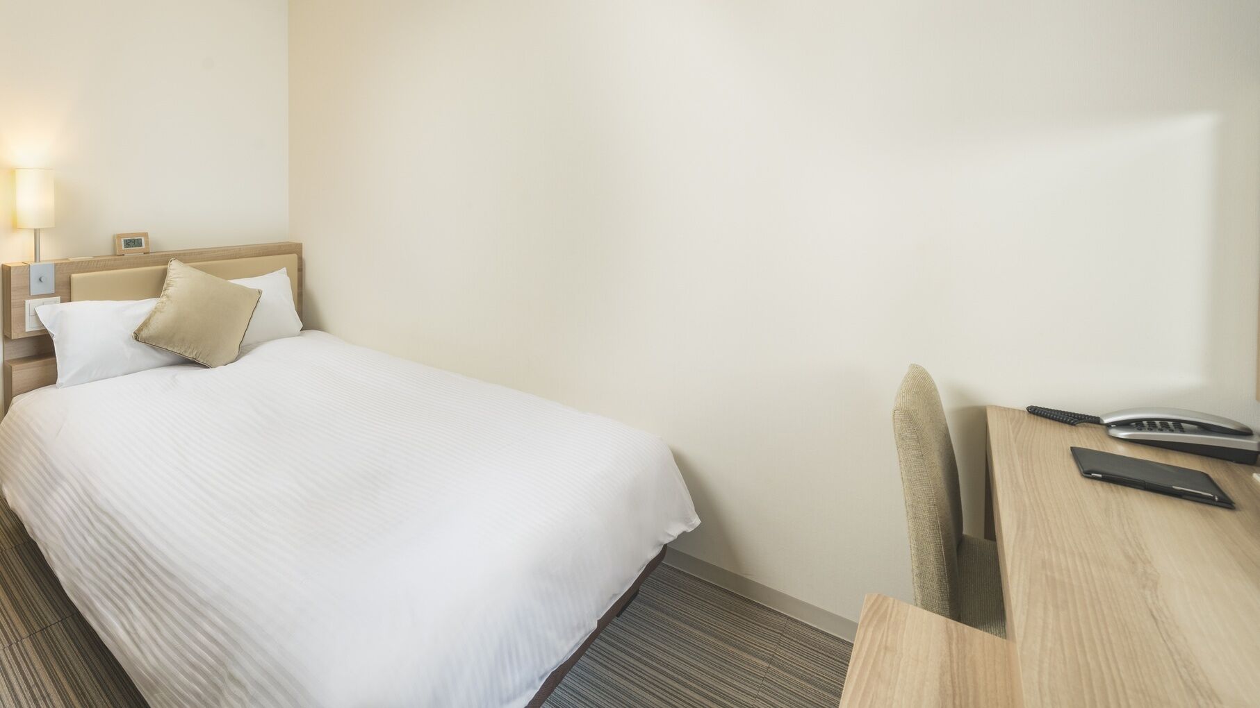 A hotel room with a bed, bedside table, lamp, and a desk with a telephone.