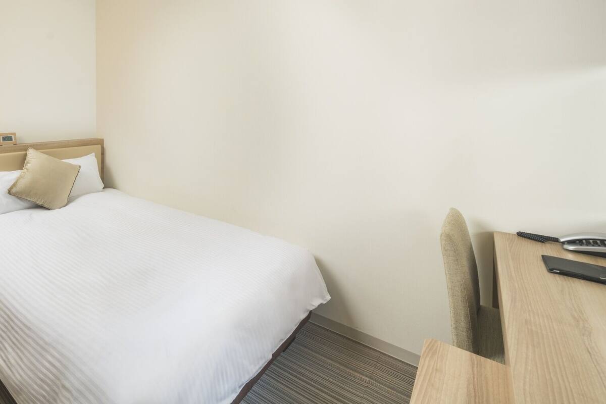 A hotel room with a bed, bedside table, lamp, and a desk with a telephone.