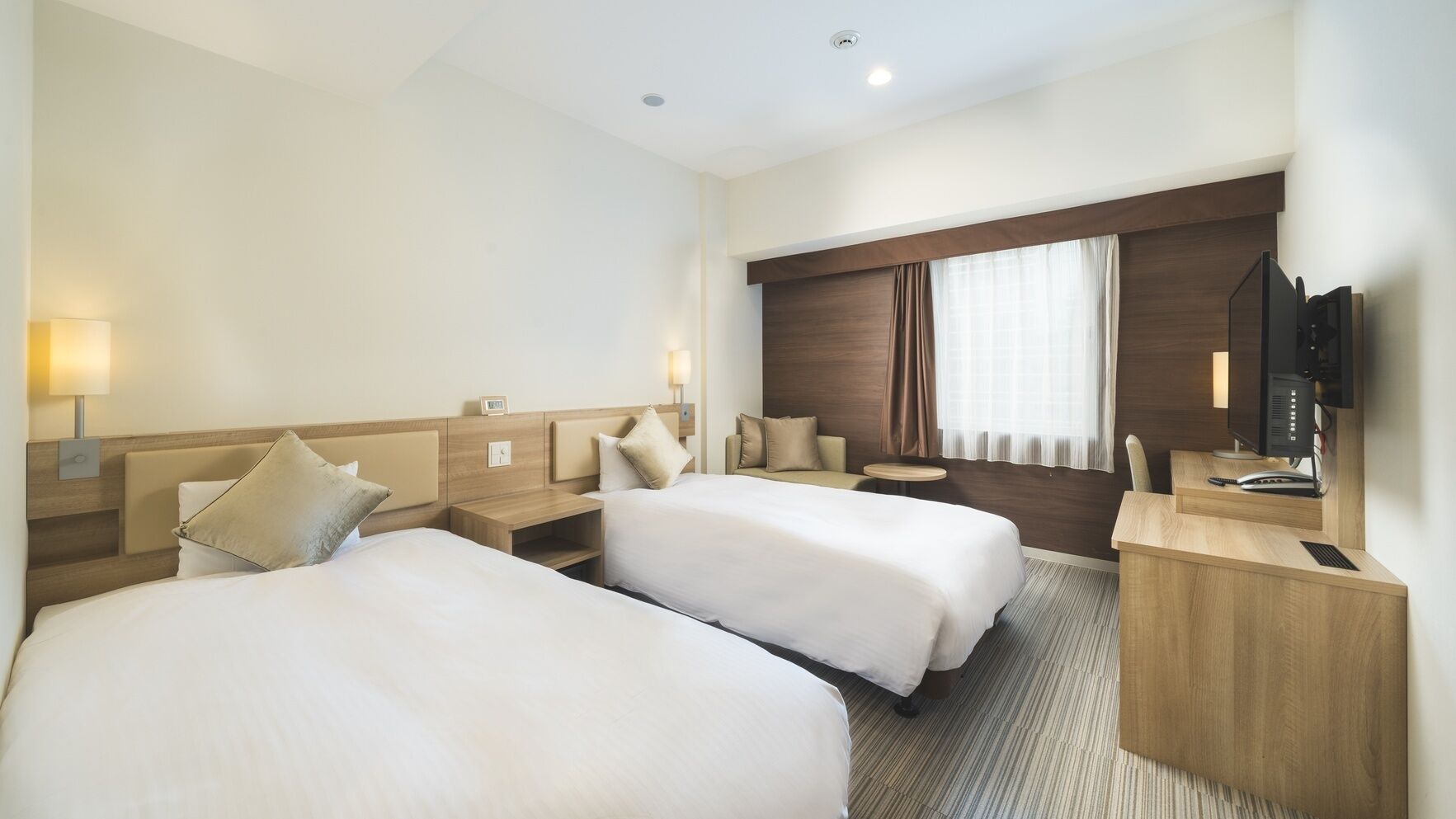A hotel room with two beds, a desk with a computer, a chair, a small table, and a window with curtains.