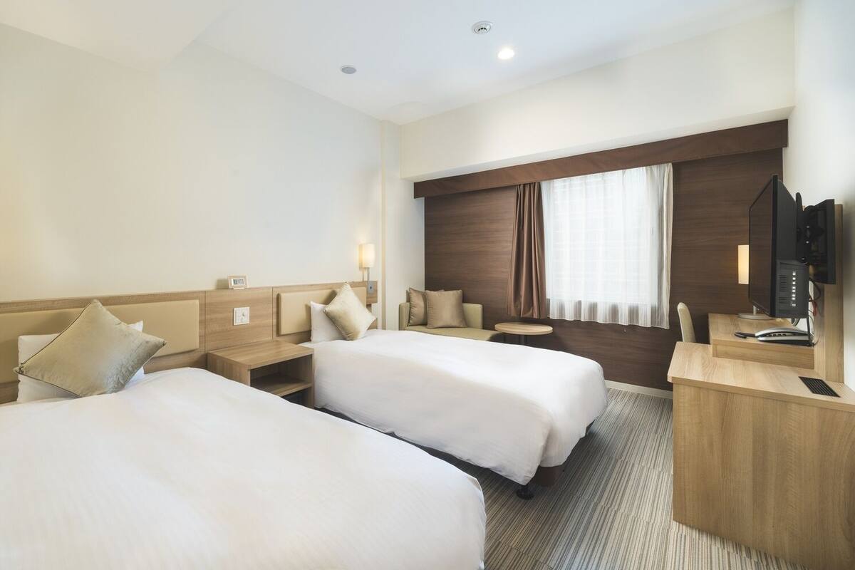 A hotel room with two beds, a desk with a computer, a chair, a small table, and a window with curtains.
