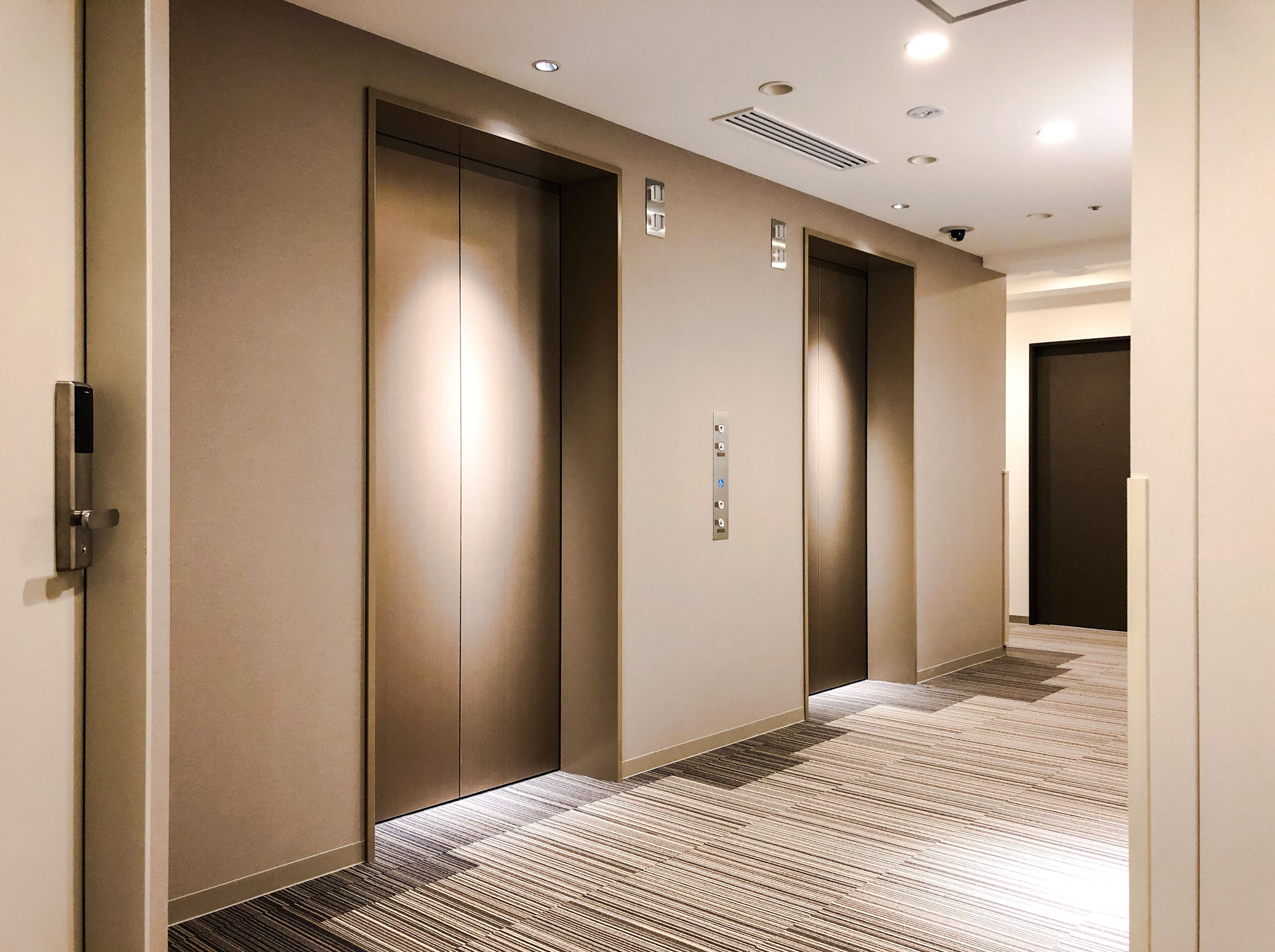 A hallway with three elevator doors, a control panel, and a patterned carpet.