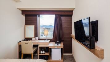 A hotel room with a bed, a desk with a computer, a TV, a mini-fridge, a coffee maker, and a window with a view of buildings and mountains.