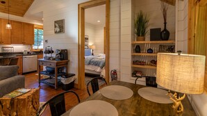A cozy room with a wooden ceiling, a bed with a blue blanket, a desk with a computer, a coffee maker, and a bookshelf with various items.