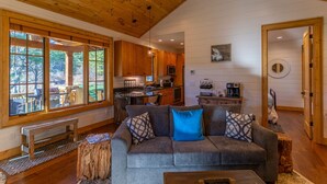 A cozy cabin interior with a wooden ceiling, a dining area with a view, a kitchen with wooden cabinets, and a living room with a gray sofa and patterned cushions.