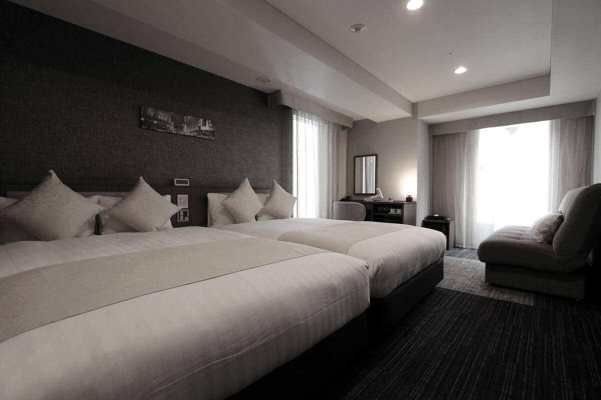 A modern hotel room with two large beds, a sofa, a desk, and a large window with curtains.