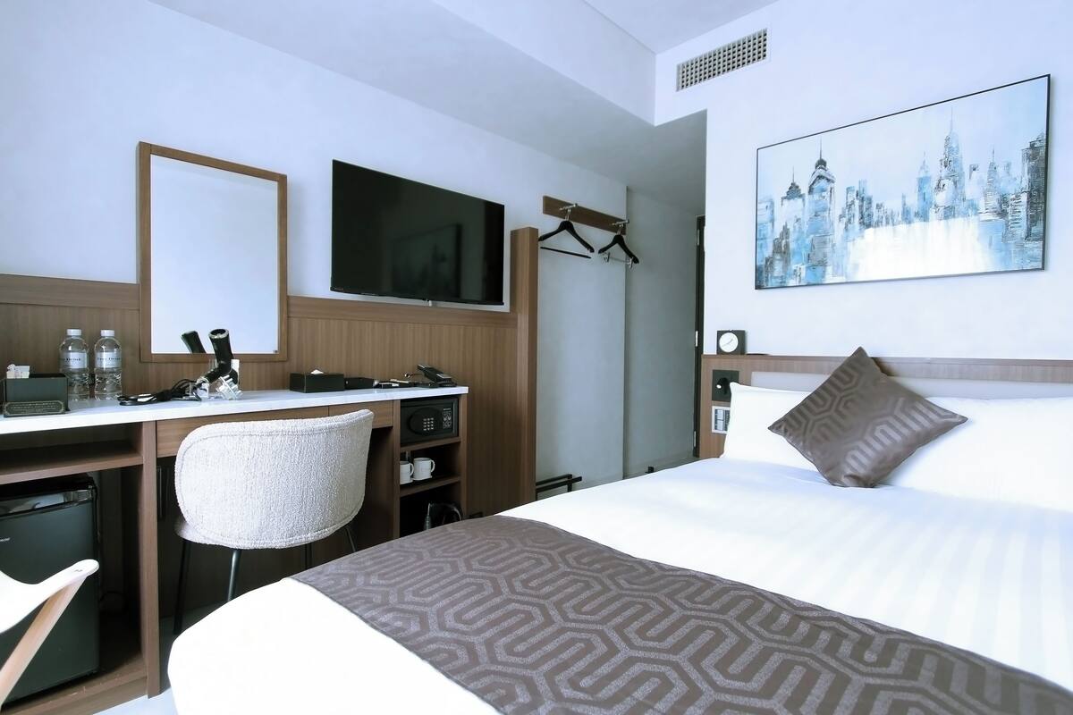 A hotel room with a bed, a desk with a chair, a television, a painting on the wall, and a coat rack.