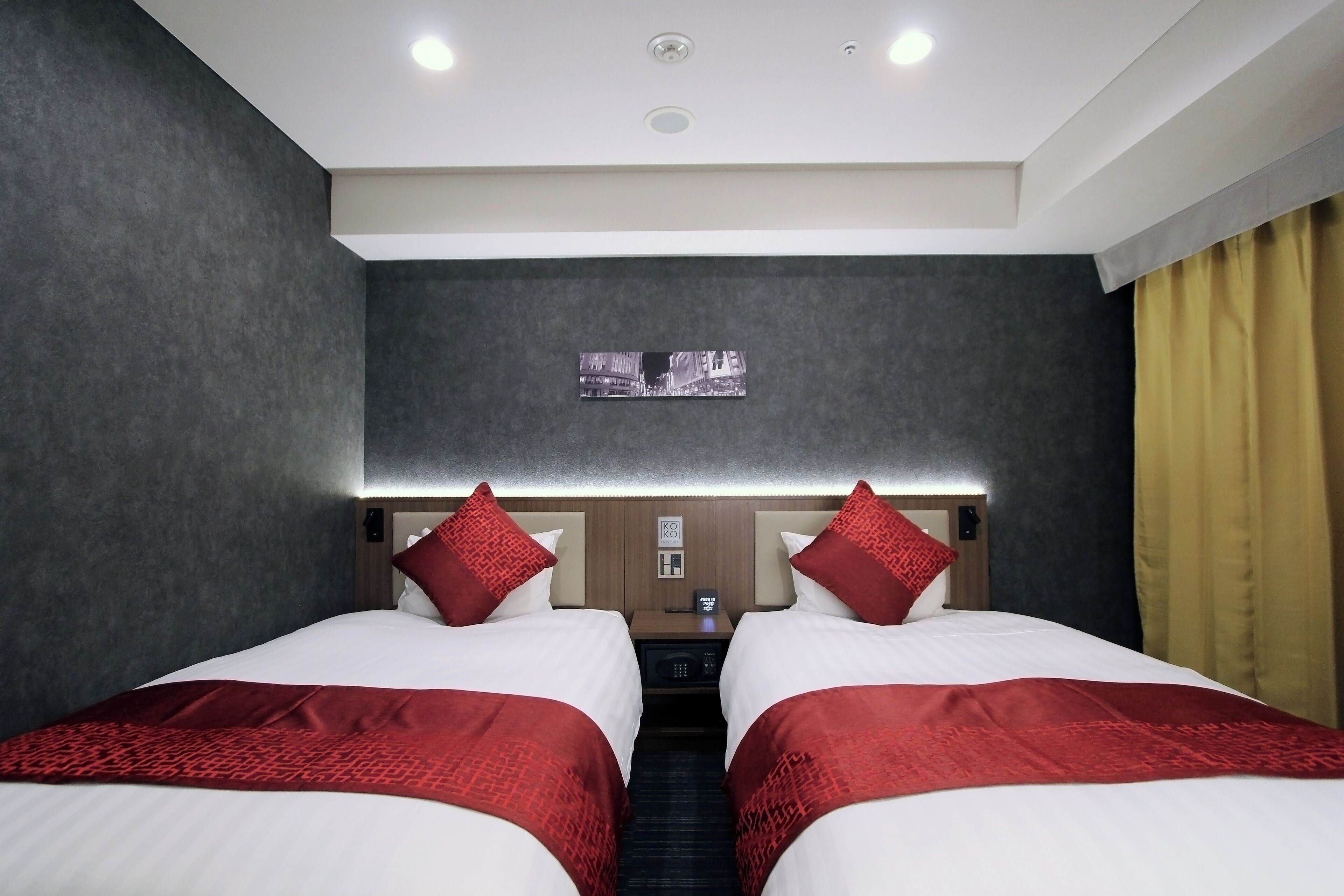 Ein Hotelzimmer mit zwei Betten, rot-weißer Bettwäsche, einem kleinen Tisch mit Telefon und einem Bild an der Wand.