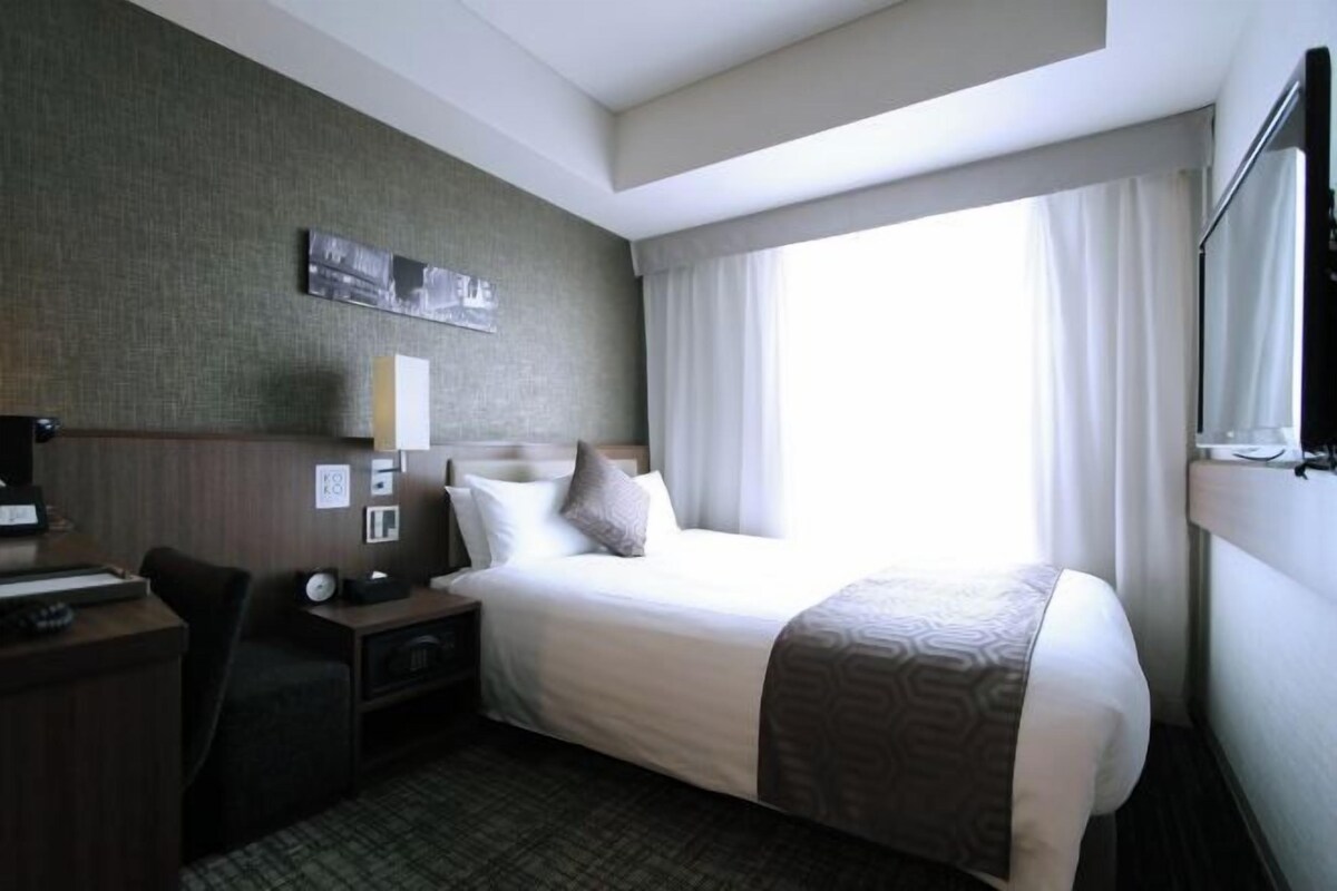 Habitación de hotel con cama, escritorio, silla, mesita de noche y ventana con cortinas.