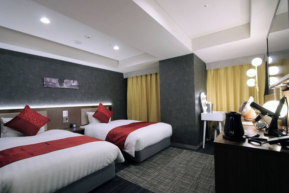 A hotel room with two beds, a desk with a lamp, a mirror, and a picture on the wall.