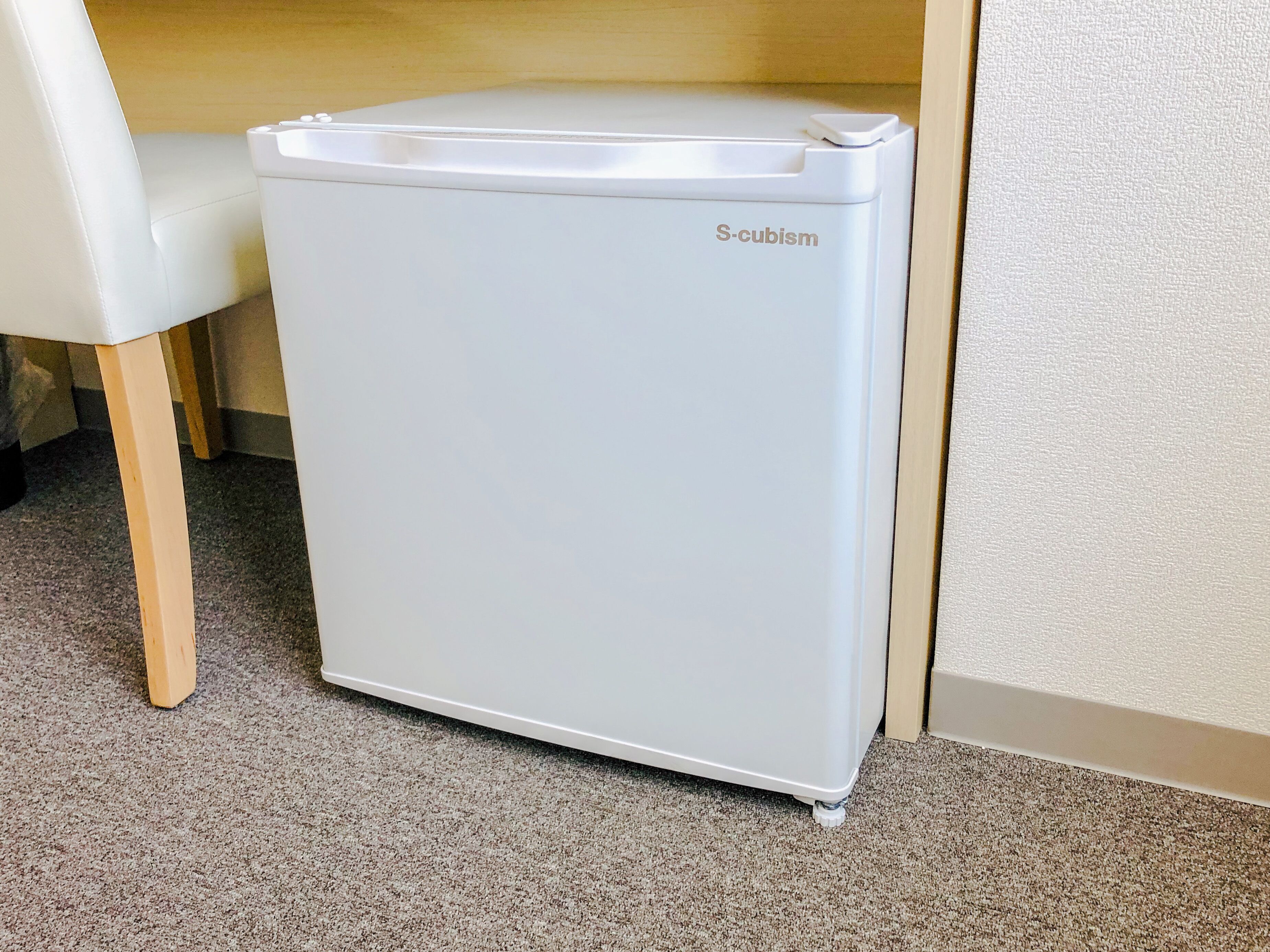 Un refrigerador blanco, de diseño cúbico, ubicado en un rincón junto a una silla de madera.