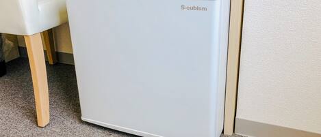 Un refrigerador blanco, de diseño cúbico, ubicado en un rincón junto a una silla de madera.