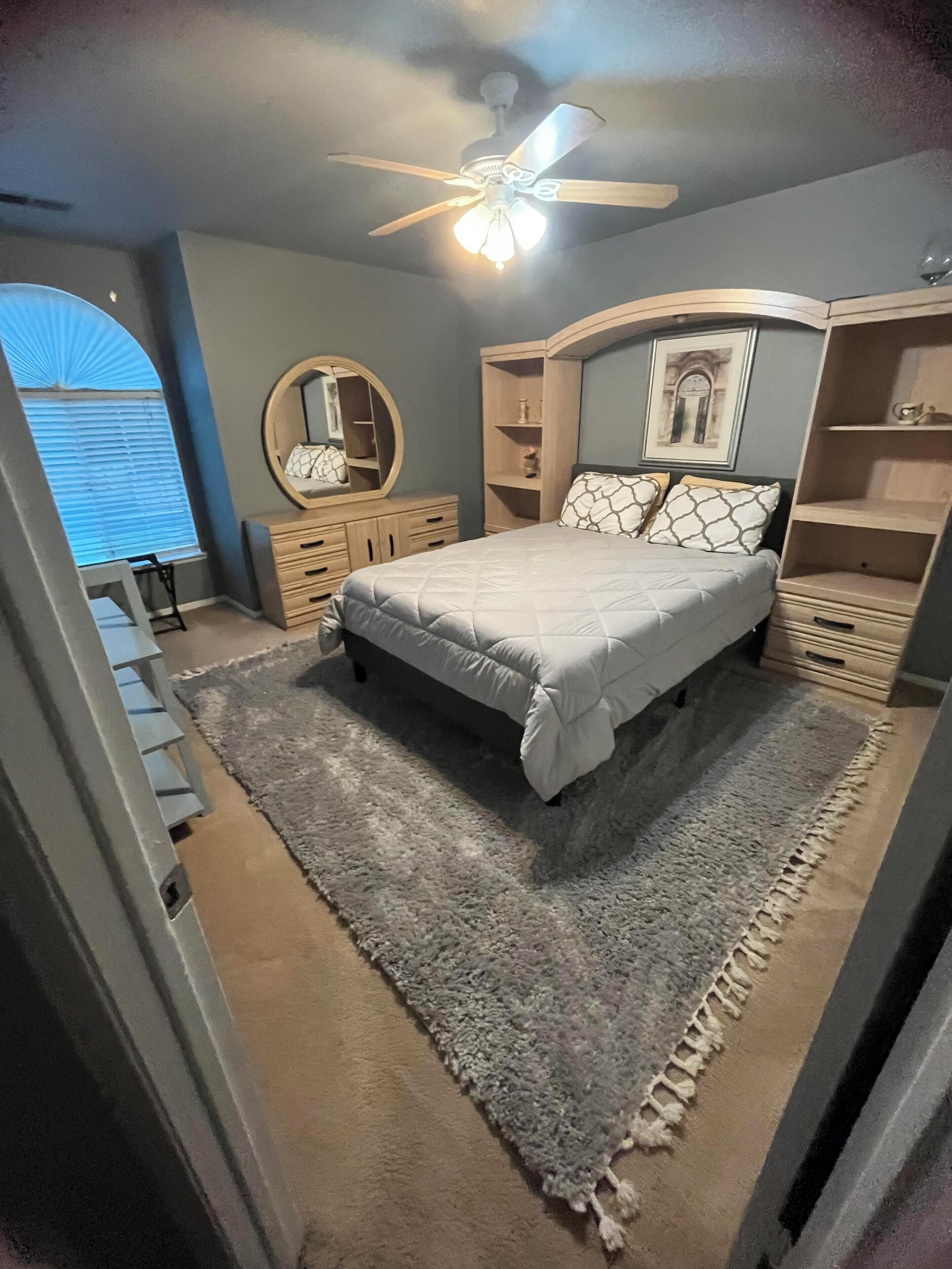 A bedroom with a bed, a ceiling fan, a dresser, a mirror, and a window with blinds.