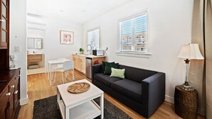 A modern living room with a dark grey sofa, a white coffee table, and a lamp. There is a kitchen area with a microwave and a sink. - Cozy Studio w/Tons of Natural Light (Coral Gables)