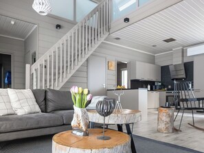 A modern living room with a grey sofa, a coffee table with a vase of tulips, a wine glass, and a rustic wooden stool. A staircase leads to an upper level. - Loma-saaga 3 by Interhome (Pudasjärvi)