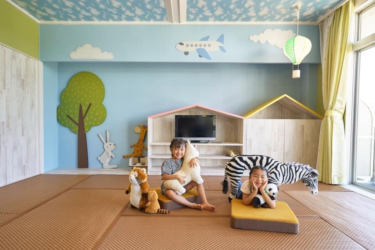 Ein Zimmer mit Tatami-Matten, einem Fernseher, einem Spielhaus und einem großen Fenster mit Vorhängen.