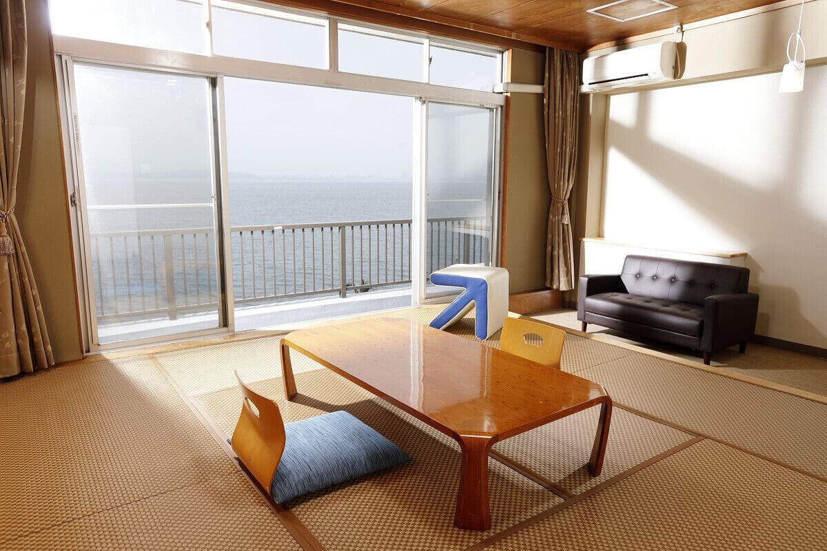 Ein Zimmer mit Tatami-Boden, einem Holztisch, einer Couch und Meerblick durch große Fenster.