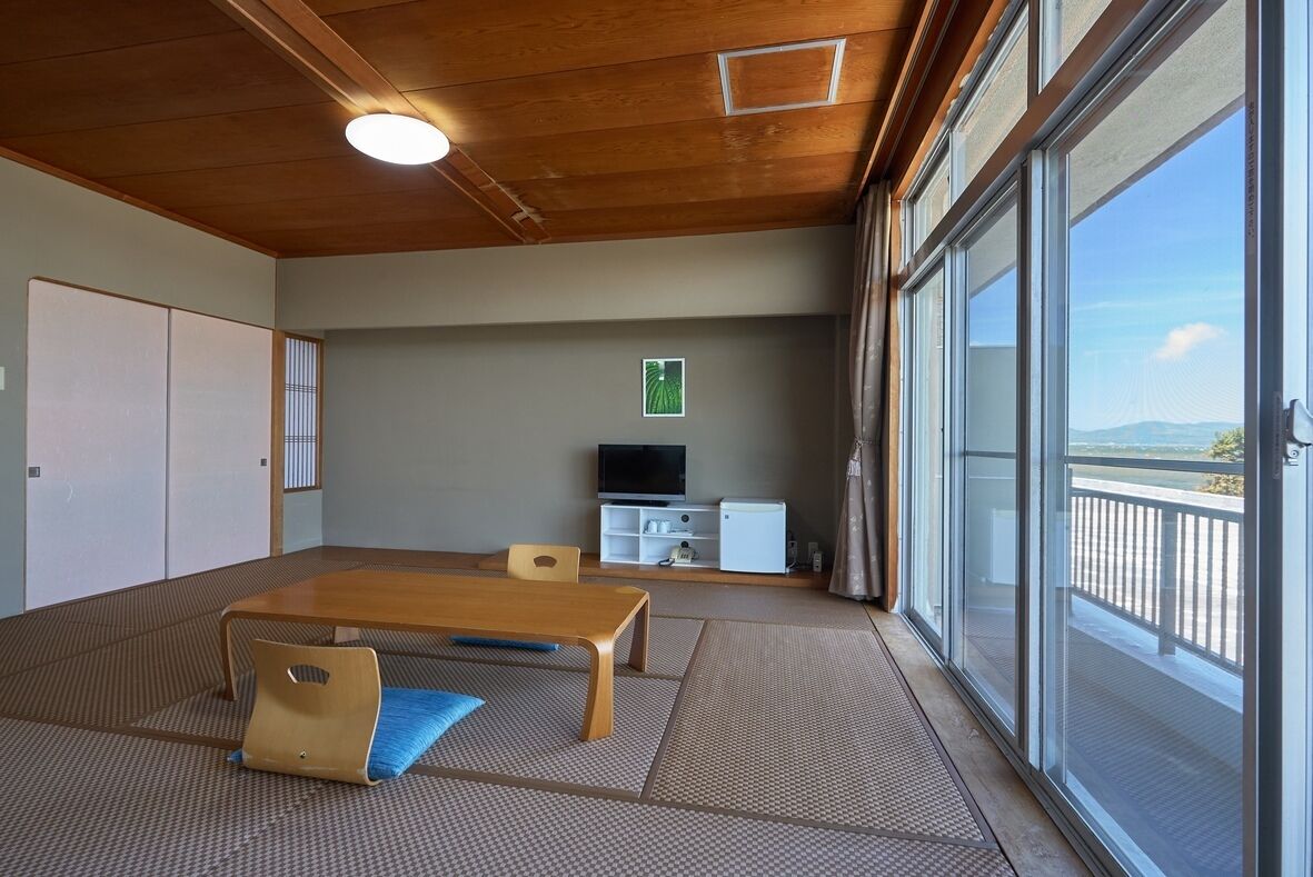 Una habitación con suelo de tatami, una mesa de madera, un televisor y una puerta corrediza de vidrio a un balcón.