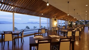 Ein Restaurant mit Holzbalkendecke und großen Fenstern mit Blick auf ein Gewässer. Die Tische sind mit weißen Tellern und Stühlen gedeckt.