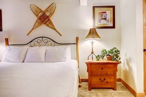 A neatly made bed with white linens, a wooden nightstand with a lamp and a plant, and a wall-mounted decorative item.