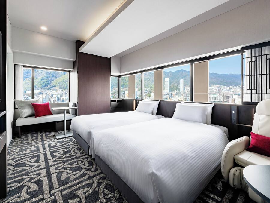 Une chambre d’hôtel moderne avec deux lits, un coin salon et de grandes fenêtres donnant sur la ville.