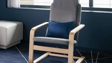 A modern chair with a wooden frame and grey and blue upholstery, positioned in a room with a dark blue carpet and a window with white curtains.