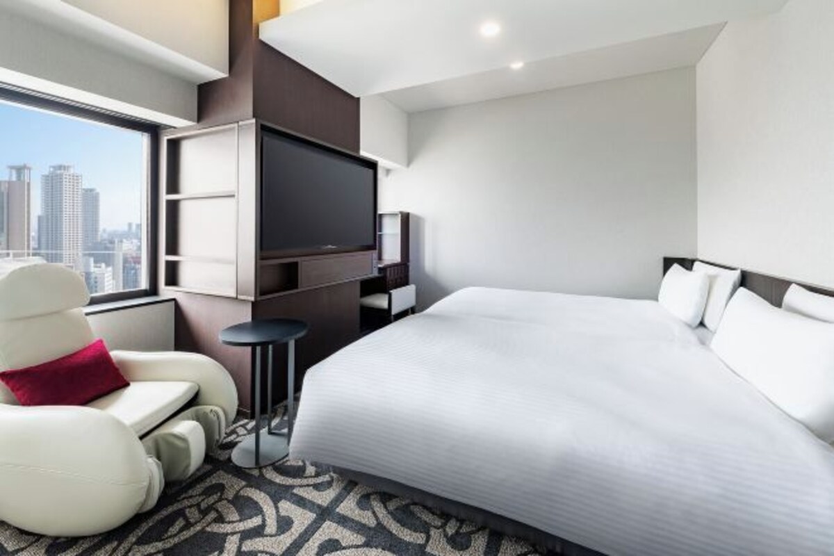 A modern hotel room with a large bed, a flat-screen TV, a white armchair, and a city view.