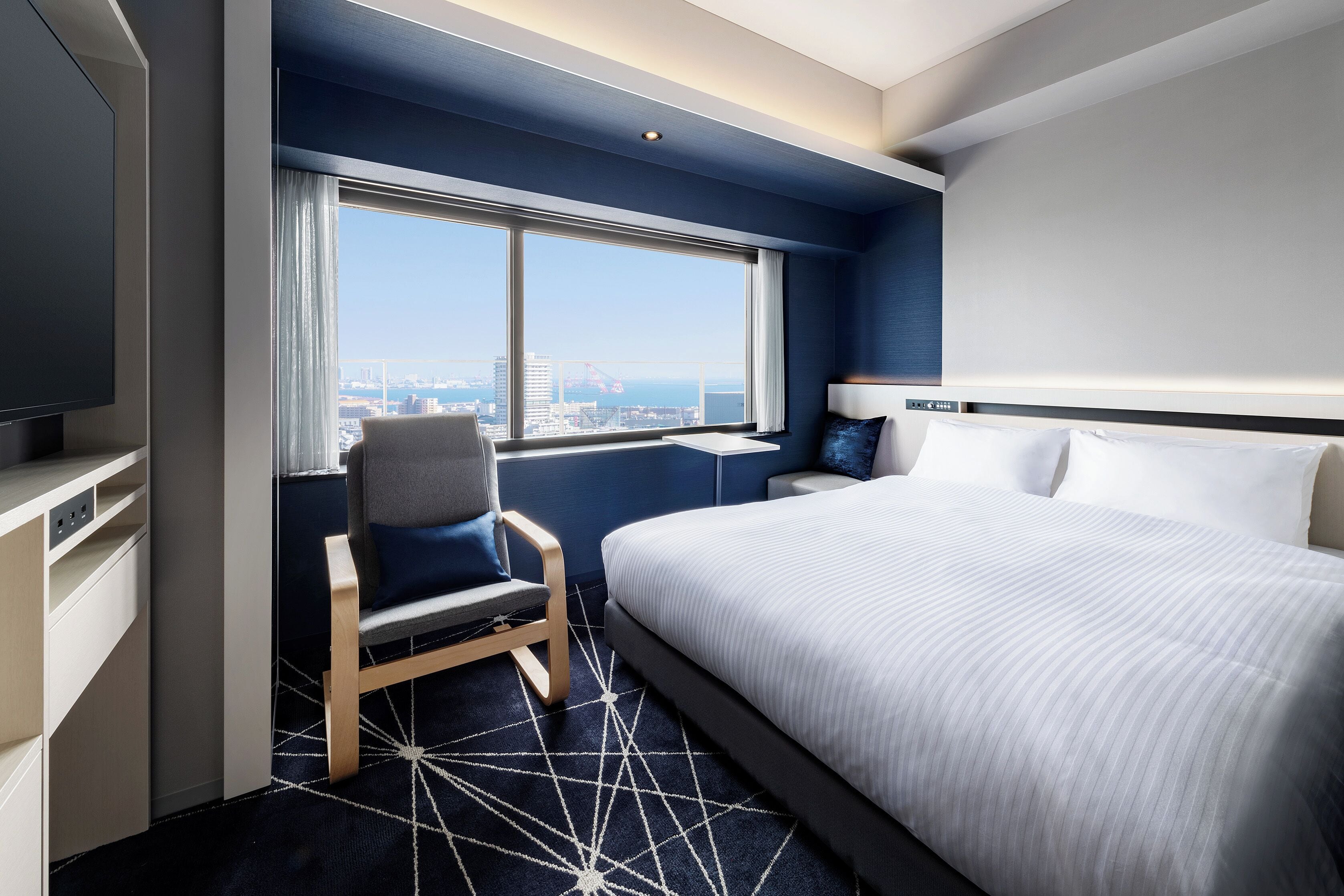 Une chambre d’hôtel moderne, dotée d’un grand lit, d’un bureau, d’une chaise et offrant une vue sur la ville par la fenêtre.