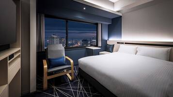 A modern hotel room with a large bed, a desk, and a chair. There is a city view through the window.
