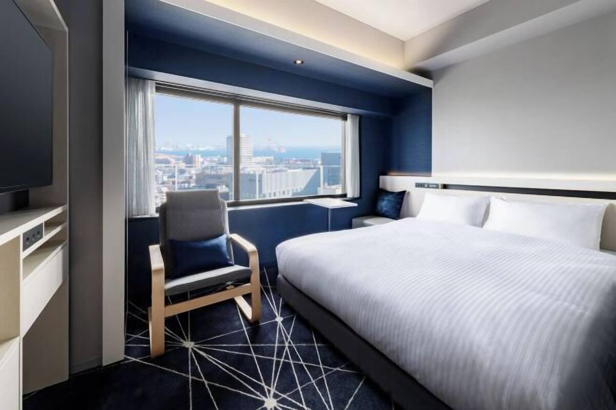 Quarto de hotel com uma cama grande, uma cadeira, uma escrivaninha e uma janela com vista para a cidade.