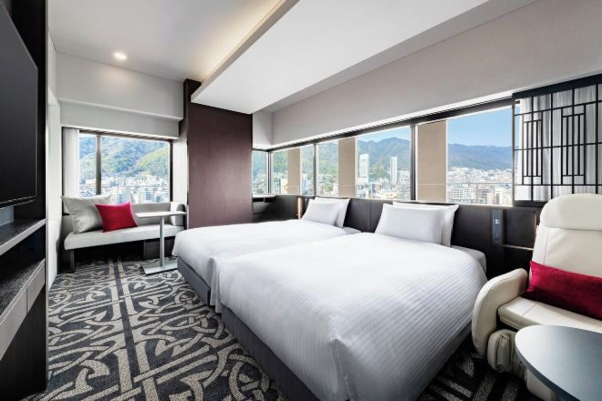 A hotel room with two beds, a seating area, and large windows with a city view.