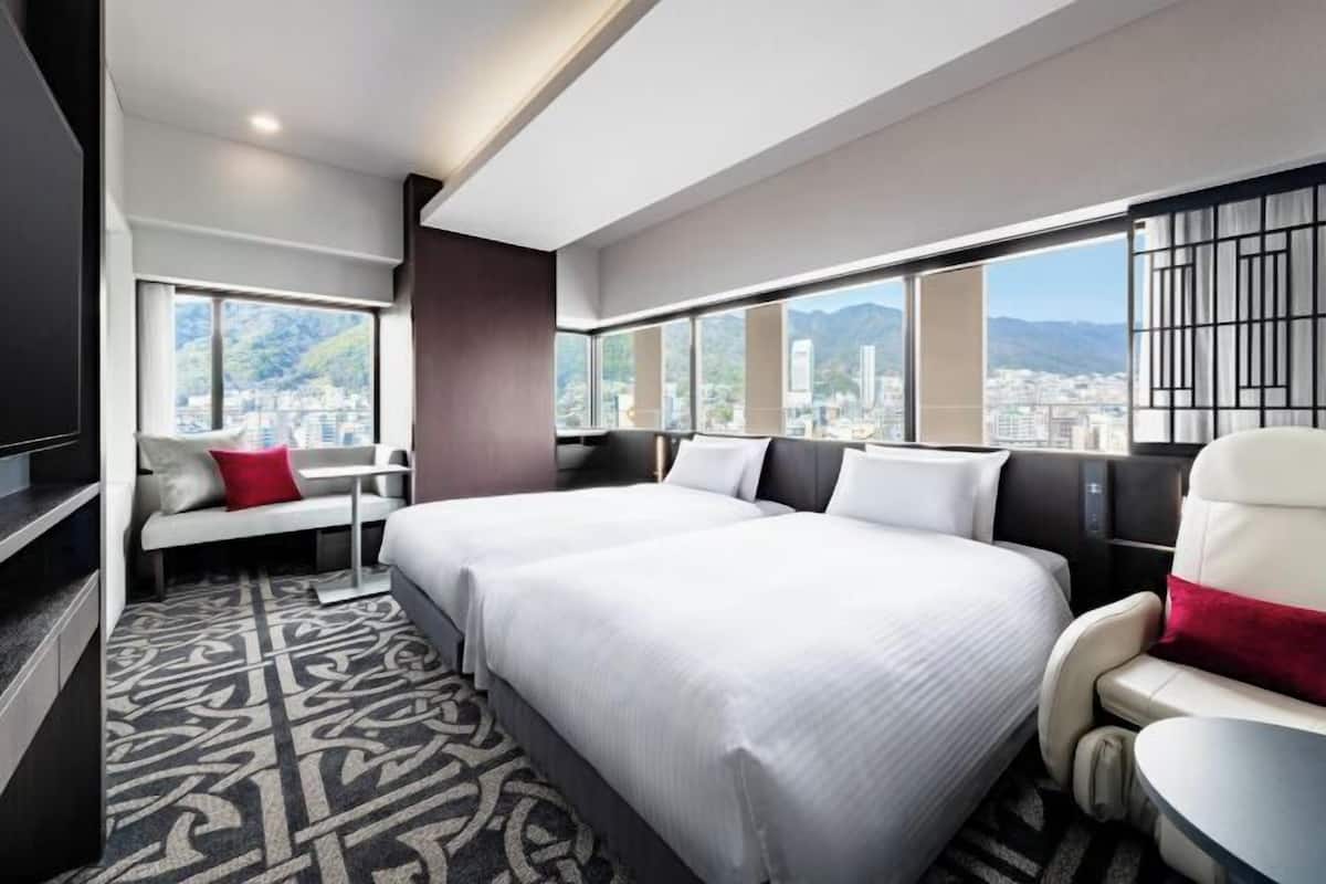 A hotel room with two beds, a seating area, and large windows with a city view.