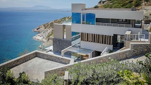 A modern coastal building with a terrace overlooking the sea, stone walls, and a clear view of the coastline. - Villa Hyperion - Luxury 7 bdrm Villa with infinity heated pool  (Rodia)