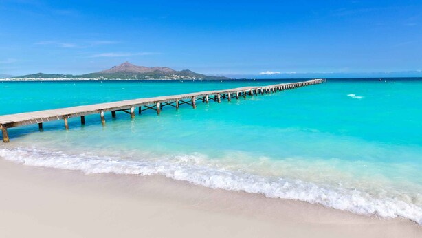 A beach with clear turquoise water, a wooden pier extending into the sea, and a mountain in the background. - Family House in Port of Alcudia | Pool, Terrace & 11 min Walk to Beach (Puerto de Alcudia)