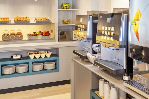 Una barra de desayuno con una máquina de café, una exhibición de frutas y platos apilados.