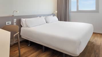 A modern hotel room with a single bed, a desk, and a chair. There is a window with curtains, a wall-mounted control panel, and a lamp.