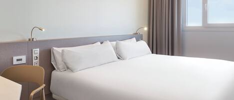 A hotel room with a bed, bedside table, chair, and a window with curtains.