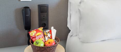 Habitación de hotel con cama, mesita de noche con una cesta de aperitivos, lámpara, teléfono y termostato.