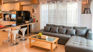 A modern living room with a grey sofa, a glass coffee table, and white bar stools. - Gala Express (Pattaya)