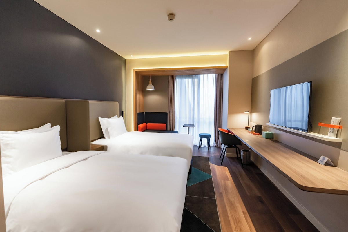 A hotel room with two beds, a desk, a television, and a seating area with a view of the outdoors.