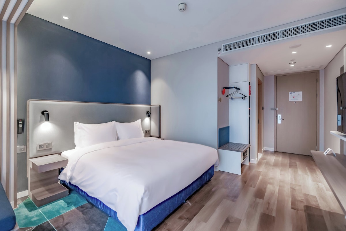 A modern hotel room with a large bed, bedside tables, a TV, and a small seating area.