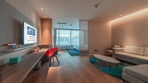 Habitación de hotel moderna con piso de madera, una silla roja, un sofá blanco, un televisor montado en la pared y un ventanal con cortinas translúcidas.