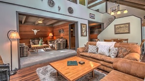 A spacious living area with a large brown leather sofa, a wooden coffee table, and a fireplace. There is a staircase leading to an upper level, and the room features a high ceiling with exposed beams. - Mountain View Townhome,| Peak 9 Access (Breckenridge)