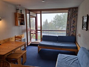 Una habitación con una mesa de madera, un banco, una cama sofá, un estante pequeño con libros, una ventana con vistas a árboles y una cortina con un estampado.