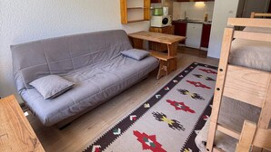 A compact living space with a sofa, a small wooden table, a kitchen area, and a patterned rug.