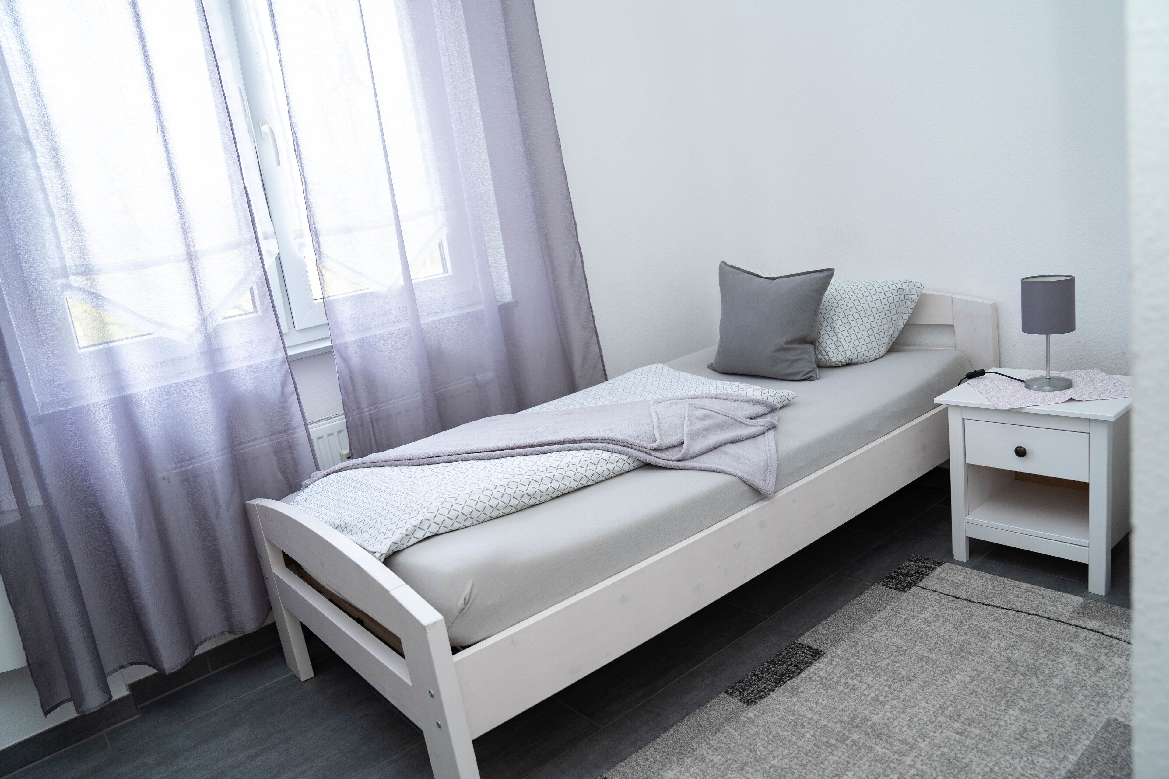A single bed with a white frame, grey bedding, and a grey pillow. There is a bedside table with a lamp and a small drawer. The room has a window with sheer curtains and a grey rug on the floor.