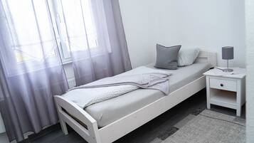 A single bed with a white frame, grey bedding, and a grey pillow. There is a bedside table with a lamp and a small drawer. The room has a window with sheer curtains and a grey rug on the floor.