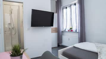 Una habitación de hotel con cama, televisión montada en la pared, ducha y ventana con cortinas.