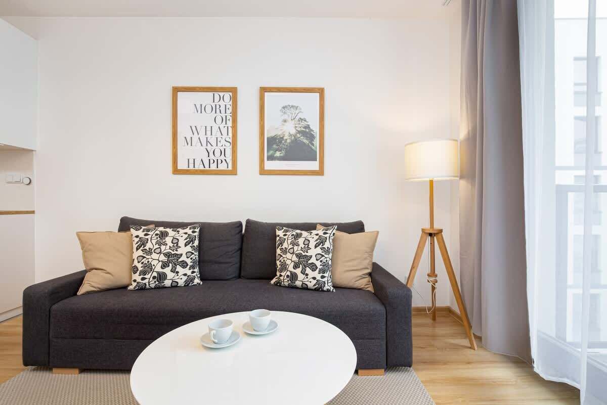 Una sala moderna con un sofá gris oscuro, dos almohadones estampados, una mesa de centro blanca con tazas, una lámpara de pie y dos carteles motivacionales enmarcados en la pared.
