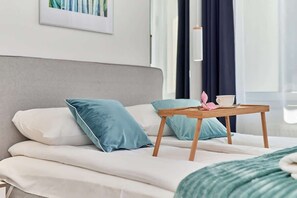 A bed with pillows, a wooden table with a cup and a pink flower, and a framed picture on the wall.