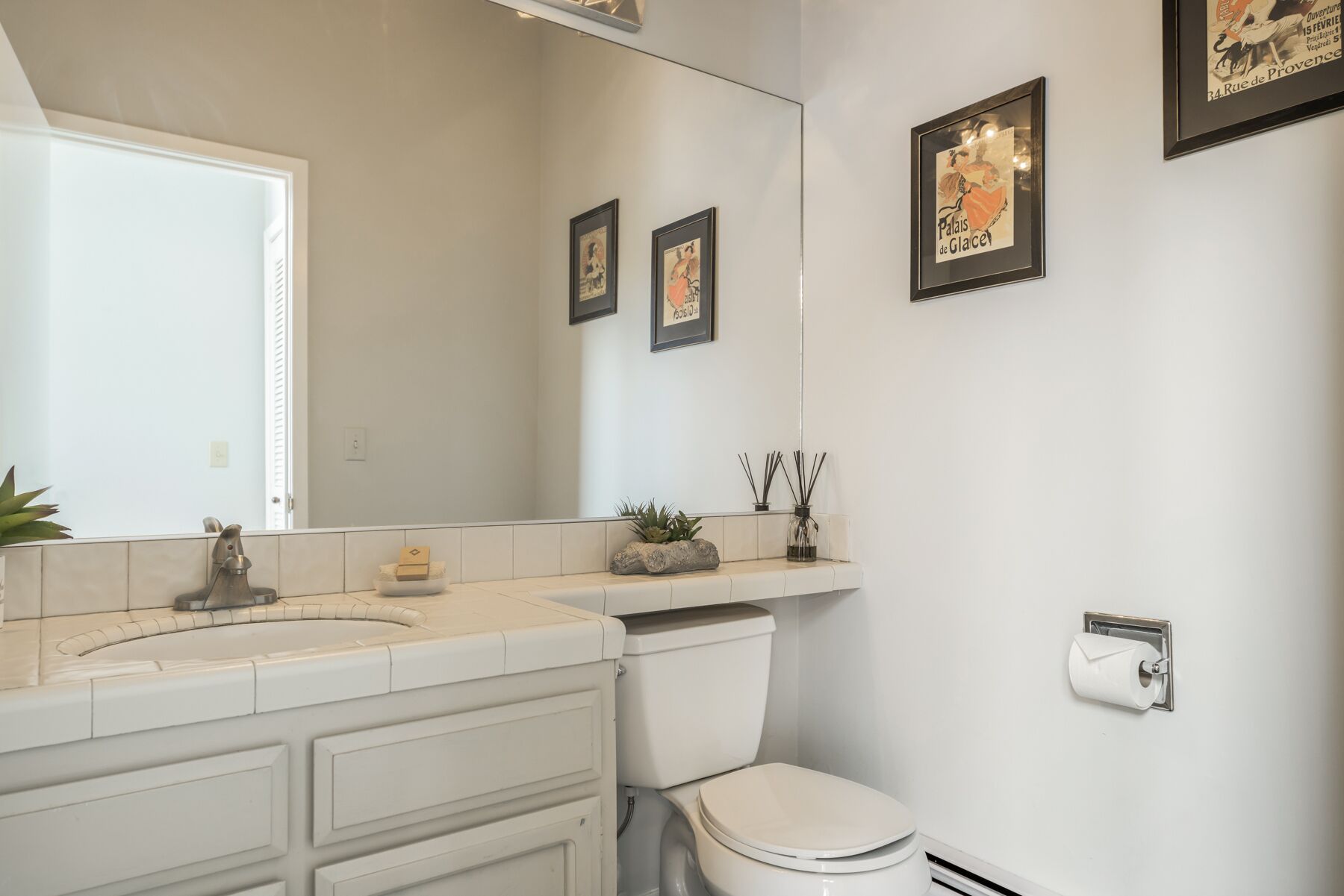 Ein Badezimmer mit weißem Waschbecken, Spiegel und Toilette. An der Wand befinden sich gerahmte Bilder und auf der Ablage steht eine kleine Pflanze.